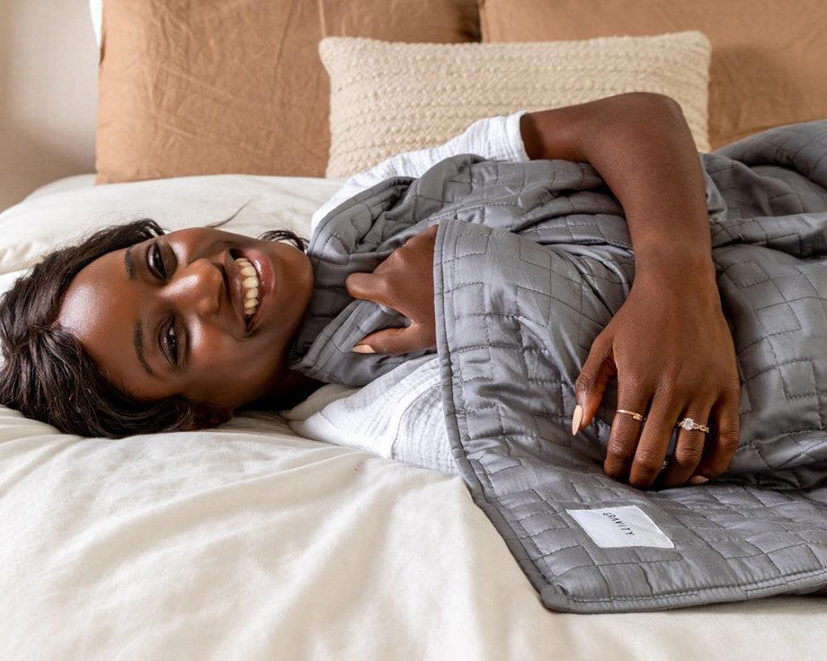 a smiling woman laying under a grey weighted blanket