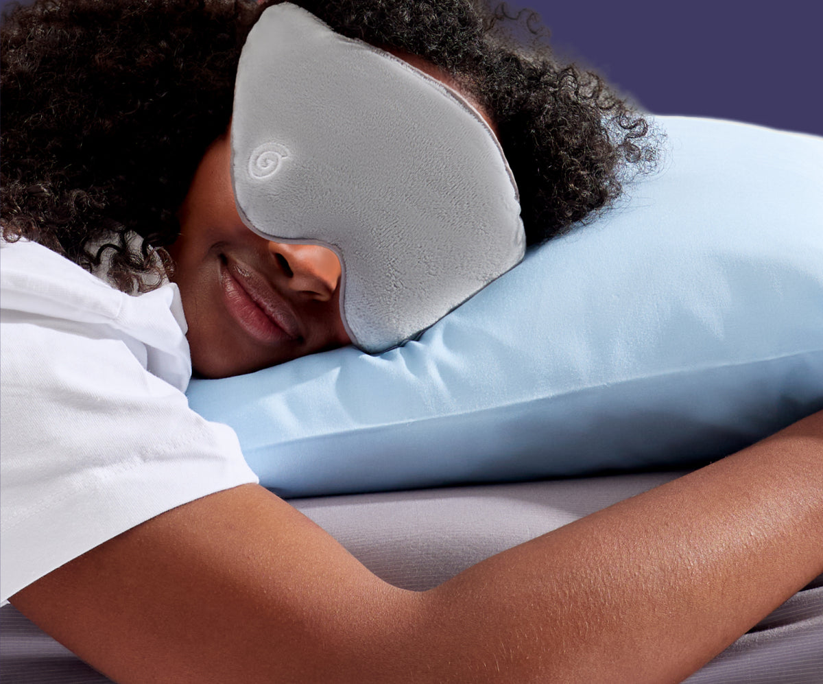 A person lying down on a pillow, wearing a grey weighted sleep mask