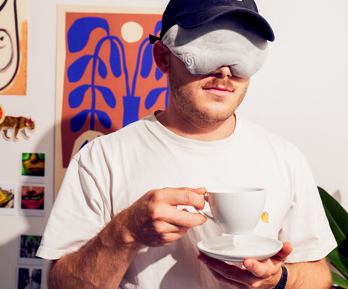 A man wearing a white t-shirt and a dark baseball cap with a grey gravity weighted sleep mask holds a white ceramic teacup and saucer. 