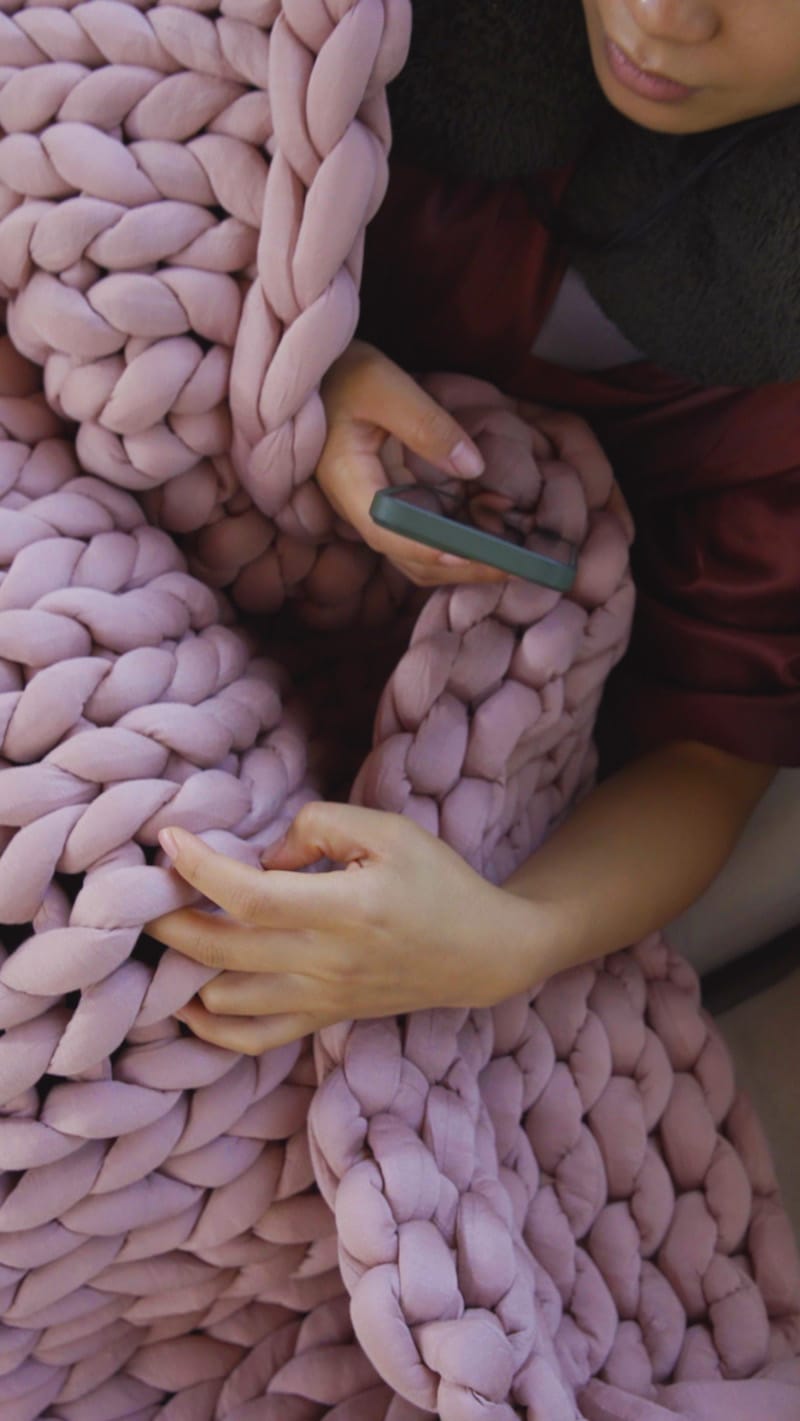 Closeup of a woman curling her fingers through a Gravity Weighted Chunky Knit Blanket while taking a photo of it on her phone.