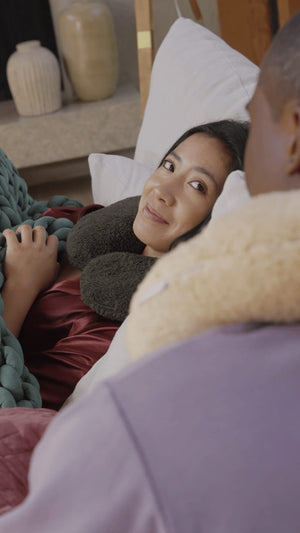 A woman laying on a sofa and wearing a Gravity Weighted Neck Pillow lovingly looking up at a man wearing a Gravity Weighted Neck Pillow standing nearby
