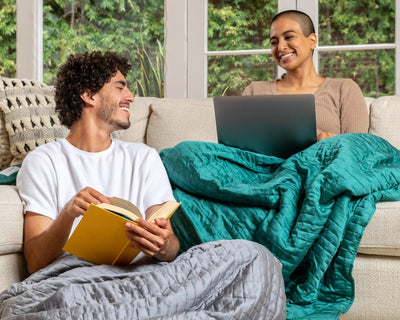 A happy couple sitting on a sofa each with a Gravity Weighted Cooling Blanket - #color_forest-green