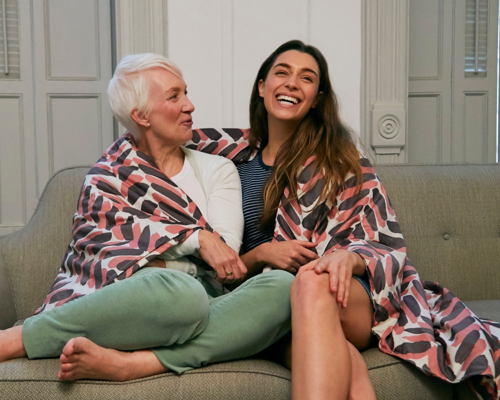  Older woman and younger woman sitting next to each other on a couch covered by a pink and brown blanket