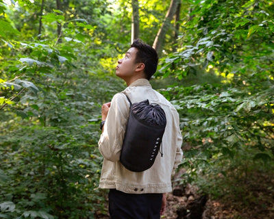 Man staring at greenery in the woods with a black travel blanket case thrown over his shoulder #color_black