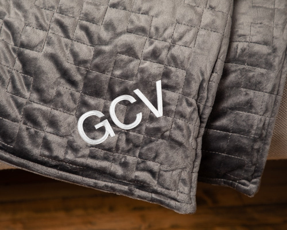 Closeup of the corner of a grey blanket draped over couch embroidered with the initials GCV - #color_grey