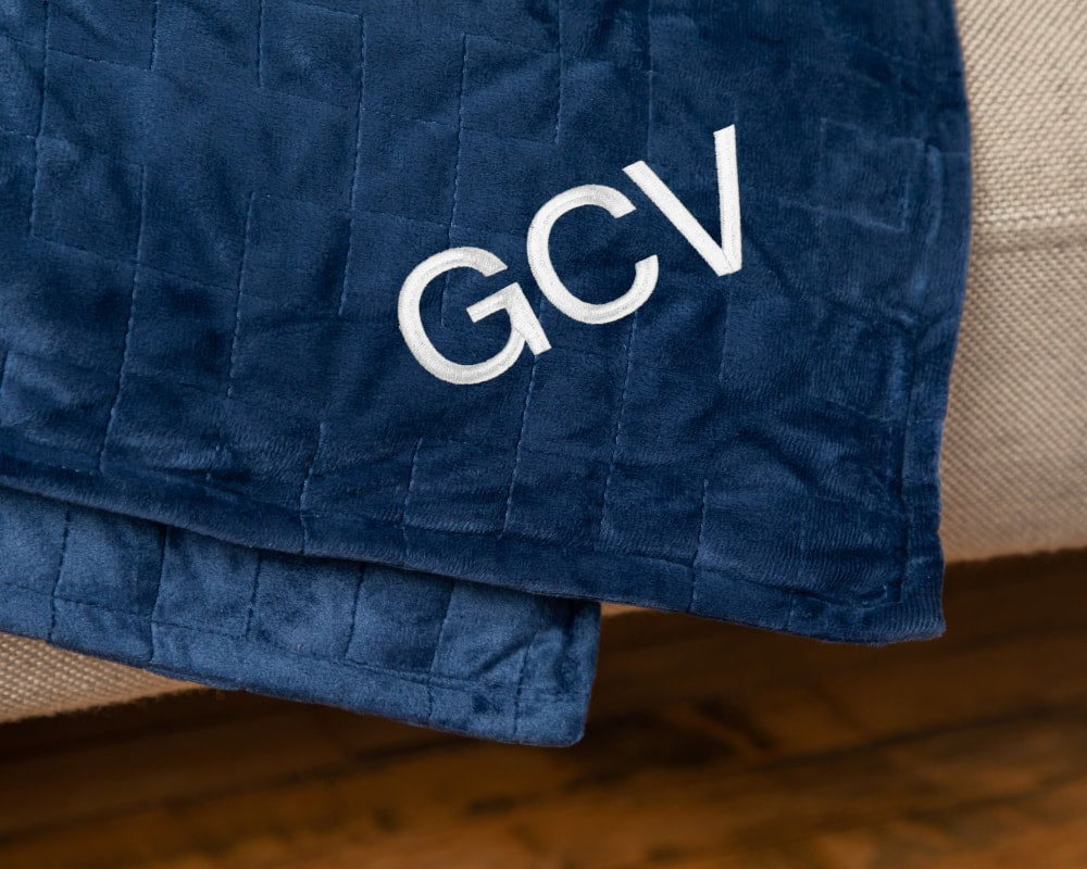 Closeup of the corner of a navy blanket draped over couch embroidered with the initials GCV - #color_navy