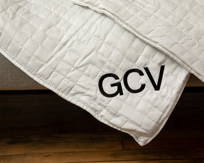 Closeup of the corner of a white blanket draped over couch embroidered with the initials GCV - #color_white