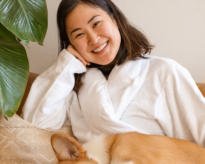 Smiling woman with a dog in her lap sitting on the couch while wearing a white terrycloth robe