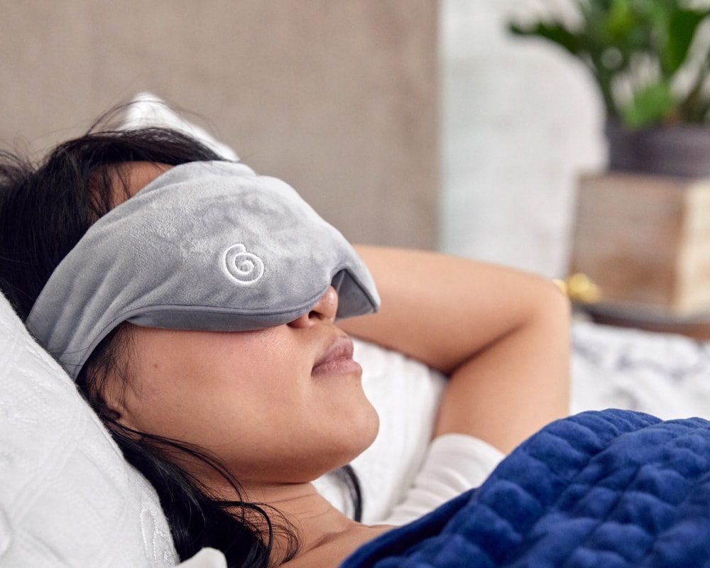 Closeup of woman's face in bed wearing a plush grey weighted eye mask
