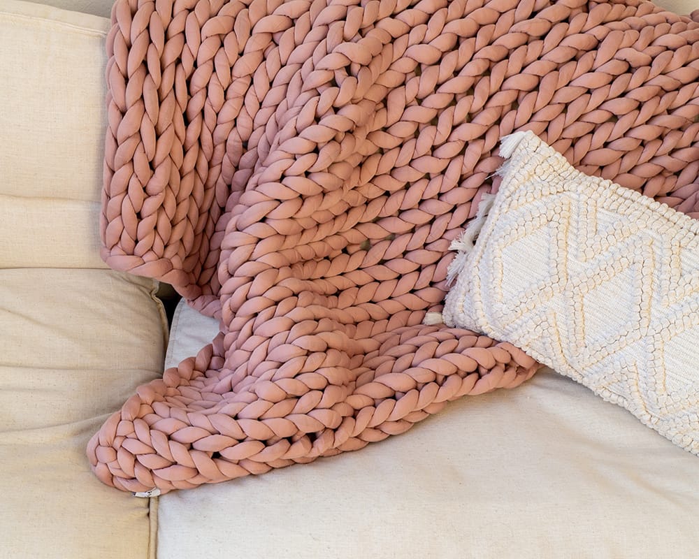 A Clay Gravity Weighted Chunky Knit Blanket draped over a sofa with a throw pillow - #color_clay
