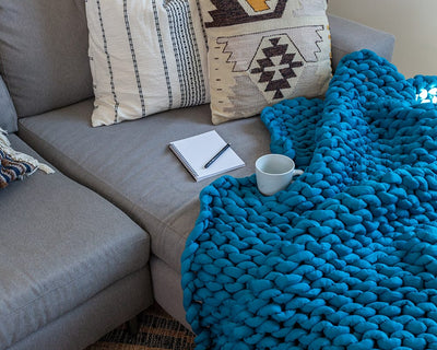 A Sapphire Blue Gravity Weighted Chunky Knit Blanket draped over a sofa with a notepad, pen, and tea cup - #color_sapphire-blue