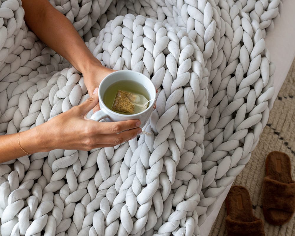 ASoft Grey Gravity Weighted Chunky Knit Blanket  on a woman's lap her hands holding a cup of tea over the blanket - #color_soft-grey